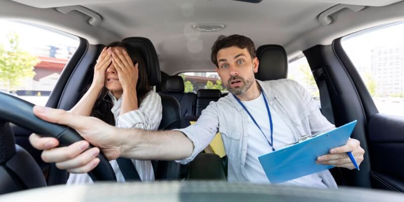 Instructor reaching over to grab steering wheel whilst girl covers her eyes
